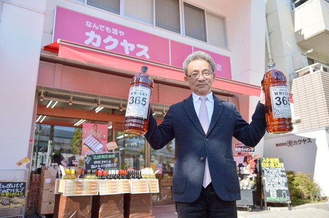 PB商品「K-Price」のウイスキー「365」を両手に掲げる社長佐藤。旗艦店の「なんでも酒やカクヤス」王子店前で